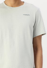 Lichtgrijs T-shirt met korte mouwen van zachte stof. Heeft een ronde halslijn en een klein logo gedrukt op de bovenste linkerborst.
