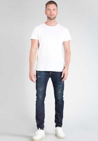 T-shirt bianco a maniche corte, jeans blu strappati e sneakers bianche. I jeans presentano sbiaditure e piccoli strappi; tessuto morbido e vestibilità classica.