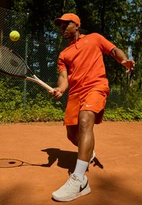 Orange polo shirt and shorts, white Nike shoes, holding a tennis racket. Tennis ball in motion on clay court, with a natural background.