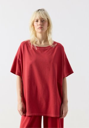 Blond osoba v voľnom červenom tričku a rovnako červených nohaviciach stojaca pred jednoduchým bielym pozadím.