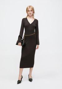 Robe tricotée côtelée marron avec un décolleté en V, de longues manches évasées et une ceinture en chaîne. Associée à un sac à main noir et des escarpins à bout pointu.