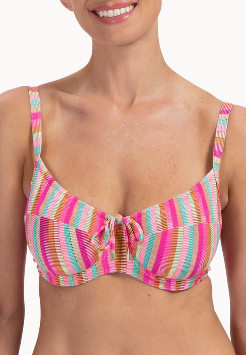 Frau trägt ein Bikini-Oberteil mit dünnen Trägern und einer vorderen Knotenbindung in pink, orange, blau und weiß gestreift, lächelt mit sichtbaren Zähnen.