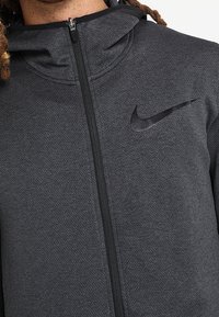 Veste à capuche noire zippée en tissu texturé, dotée d'un logo Nike discret sur le côté gauche de la poitrine, d'un col montant et d'une fermeture éclair à l'avant.