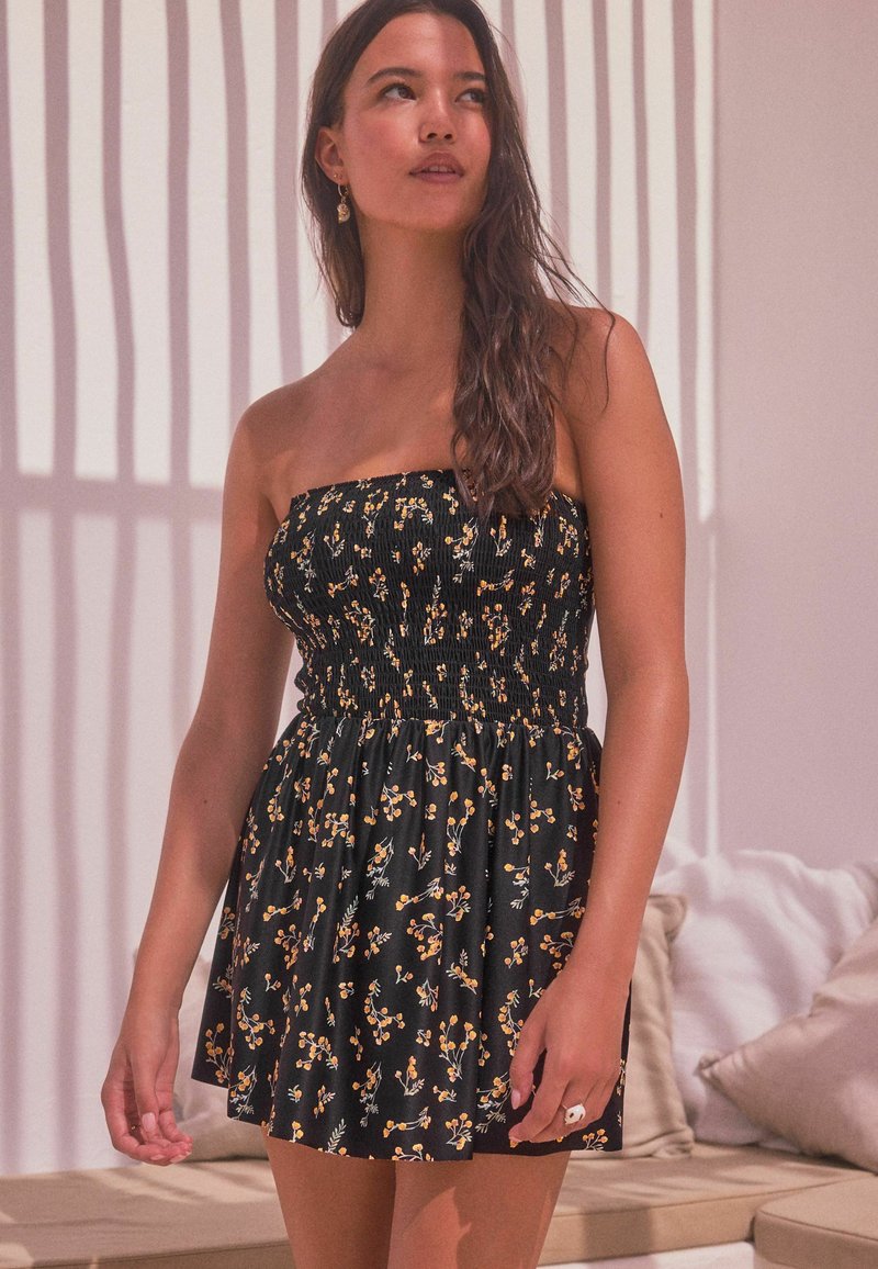 Vestido negro sin mangas con un cuerpo fruncido, que presenta un patrón floral en amarillo y verde. Falda corta, hecha de un tejido ligero.