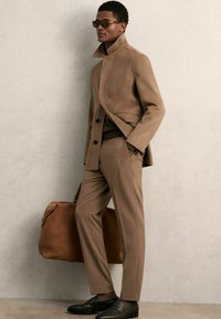 Brauner Wollmischmantel mit hohem Kragen, kombiniert mit passenden taillierten Hose und dunklen Leder Slippern. Begleitet von einer braunen Handtasche.