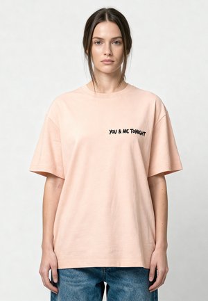 Femme aux cheveux bruns portant un t-shirt oversize rose pâle avec le texte « YOU & ME TONIGHT », debout sur un fond clair uni.