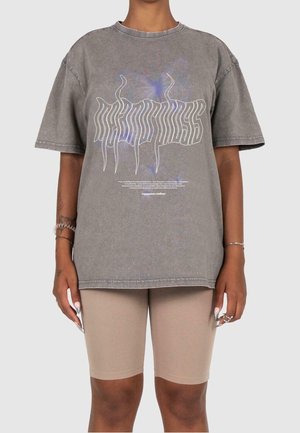 Person iført en løs grå grafisk t-shirt med forvrænget tekst og et svagt sommerfugledesign, kombineret med beige biker-shorts.