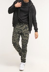Zielone spodnie bojówki w kamuflażu z czarnymi wzorami, z kieszeniami i wąskim krojem. Do tego czarna bluza z kapturem i białe trampki.