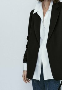 Blazer nero su misura con una texture liscia, rever a punta e maniche lunghe indossato sopra una camicia bianca a bottoni e jeans blu.