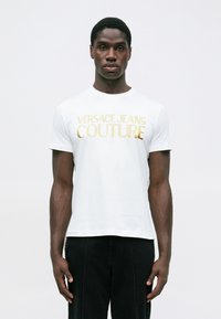 Bijela pamučna majica s zlatnim logotipom "VERSACE JEANS COUTURE" preko prsnog dijela, okrugli izrez i kratki rukavi.