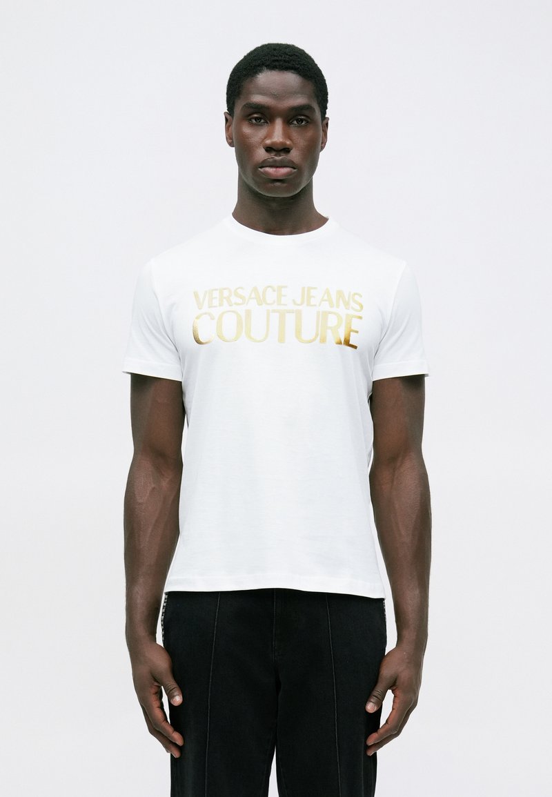 Bijela pamučna majica s zlatnim logotipom "VERSACE JEANS COUTURE" preko prsnog dijela, okrugli izrez i kratki rukavi.