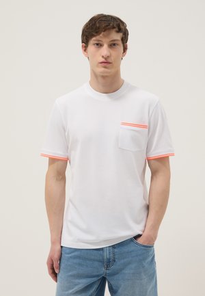 Giovane uomo che indossa una maglietta bianca con bordo arancione sulla tasca e sulle maniche, in piedi con una mano in tasca su uno sfondo semplice.