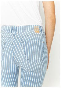 Světle modré denimové džíny s vertikálními bílými pruhy, které nosí osoba v bílé košili, se zobrazenými zadními kapsami a štítkem na pase.