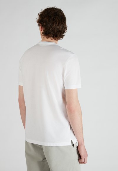 T-shirt bianco a maniche corte con scollatura a girocollo, texture morbida e spacchi laterali all'orlo, indossato con pantaloni grigi chiari.