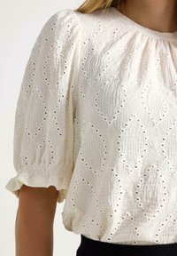 Hvid tekstureret bluse med pufærmer og diamantformede hulmønstre båret af en person med lys hud og blondt hår.
