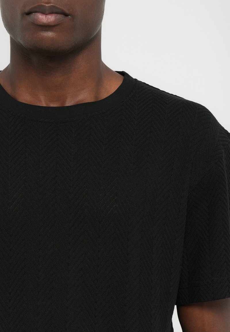 Black textured knit T-shirt featuring a herringbone pattern, round neckline, and short sleeves. Material appears lightweight and breathable.