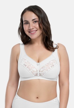 Uśmiechnięta kobieta ubrana w biały biustonosz z koronką i dopasowaną bieliznę, stojąca na jednolitym jasnym tle.