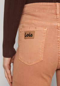 Traperice breskvaste boje s teksturiranim materijalom. Sadrže smešnu kožnu oznaku s "Lois" logotipom na zadnjem džepu. Slim fit dizajn.