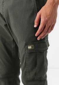 Grüne Cargo-Hosen aus leichtem Stoff, mit seitlichen Taschen und einem "TOMMY JEANS"-Logopatch am linken Oberschenkel.
