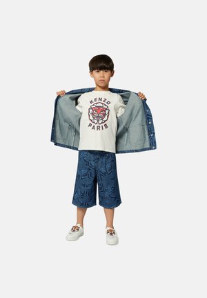 Enfant portant une veste en denim, un t-shirt blanc avec un motif de tigre, et un short bleu à motifs. Baskets avec un design de tigre sur les côtés.
