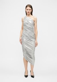 Γυναίκα με ασημί μεταλλικό midi φόρεμα ενός ώμου με ραφές, συνδυασμένο με μαύρες μυτερές γόβες, στέκεται μπροστά σε απλό φόντο.