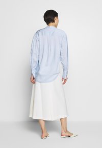 Camicia a maniche lunghe azzurro chiaro con maniche a palloncino, dettaglio a piega sul retro e polsini con bottone, abbinata a una gonna bianca a trapezio e scarpe basse a punta.