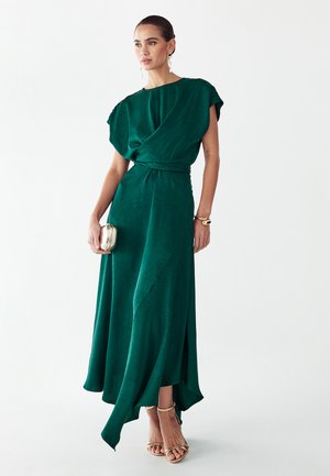 Frau trägt ein ärmelloses, dunkelgrünes asymmetrisches Kleid, hält eine goldene Clutch, mit goldenen Sandalen und goldenem Schmuck vor weißem Hintergrund.