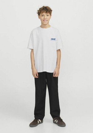 Vit bomullströja med blå "OMC"-logotyp på vänster bröst, kombinerad med svarta, löst sittande byxor och bruna sneakers med vita ränder.