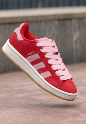 Baskets Adidas Campus rouges avec des rayures roses et des lacets, semelle blanche et semelle extérieure en gomme, positionnées en l'air au-dessus du sol en béton.