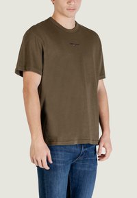 T-shirt in cotone verde oliva con maniche corte, scollo rotondo e piccolo logo ricamato sul petto, abbinata a jeans in denim blu.