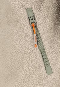 Hellbeige Fleece-Stoff mit einem grünen Reißverschluss und einem orangefarbenen Zipper. Der Reißverschluss ist teilweise geöffnet und zeigt ein strukturiertes Innenleben.