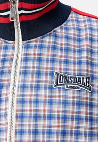 Geruite jas met blauwe, rode en witte patronen en een marineblauwe kraag. Ritssluiting aan de voorkant, geborduurd "Lonsdale London" logo op de borst.