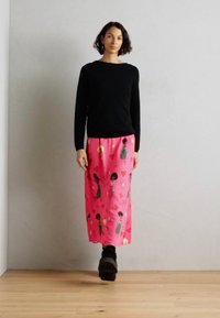 Svart genser, rosa ankel-langt skjørt med illustrerte figurer og hjerter, glatt stoff, tettsittende liv, står på tregulv mot grå vegg.
