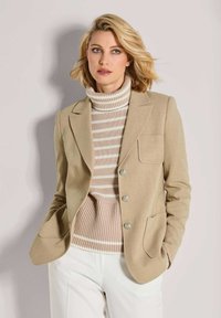 Vrouw in een beige blazer over een beige en witte gestreepte turtleneck trui en witte broek, staand tegen een lichte achtergrond.