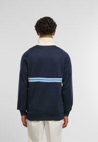 Mornarsko modra pulover s belim ovratnikom in tremi svetlo modrimi vodoravnimi črtami čez hrbet. Mehkoba tkanine in sproščen kroj.