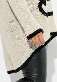 Beige gebreide trui met zwarte details, met een losse snit en een oversized pasvorm. De textuur is open geweven, wat de zachte, ademende stof benadrukt.
