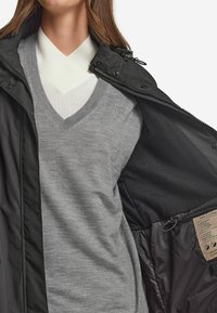 Svart ytterjacka med slät yta, lager ovanpå en ljusgrå v-ringad tröja. Inuti visas en matt svart foder och en etikett.