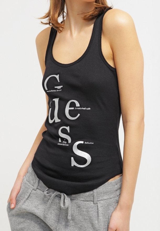 Kobieta w dopasowanej czarnej koszulce na ramiączkach z pionowym napisanym logo "Guess" oraz w szarych dresach z wiązanym pasem.