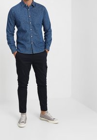 Camisa de mezclilla azul de botones con pequeños patrones, combinada con pantalones oscuros de corte ajustado y zapatillas ligeras de color gris. Las telas parecen suaves y informales.