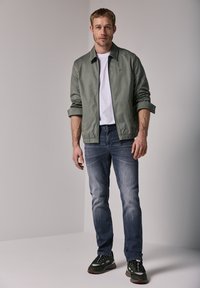 Světle zelená bomber bunda s bílým tričkem pod ní, doplněná o vybledlé modré džíny a zelené tenisky s černými akcenty.