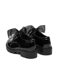Zapatos de charol negro con suela gruesa, con un gran lazo negro translúcido atado en la parte trasera y cierre plano con cordones.