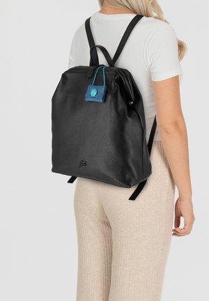 Sac à dos en cuir noir à texture granuleuse avec des bretelles ajustables et une étiquette bleue. Il possède une forme lisse et structurée avec un logo subtil en bas.