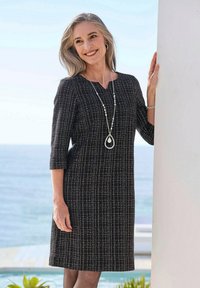Robe texturée noir et blanc avec des manches trois-quarts, coupée au genou et décolleté en V. Portée avec un long collier pendentif en argent.