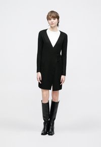 Svart strikket omslagskjole med lange ermer, V-hals og midjebånd, kombinert med knelange svarte skinnstøvler.