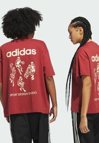 Červené oversized tričko, které na zádech má bílé logo adidas a sportovní ilustrace. Materiál vypadá měkce a má volný střih.