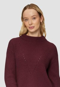 Burgunderfarbener gerippter Pullover mit hohem Kragen, der ein V-förmiges Design und kleine Aussparungen über der Brust aufweist, hergestellt aus einem weichen Stoff.