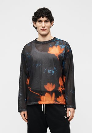 Joven con cabello rizado que lleva una camisa negra de manga larga de malla con patrones florales naranjas y azules, y pantalones negros con cordones blancos.