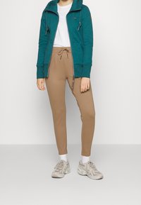 Teal rits hoodie met voorzakken, gecombineerd met beige tapered joggingbroek. Schoenen zijn lichtgrijze sneakers met een patroon.