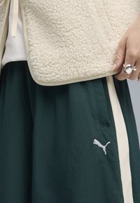 Krämfärgad, strukturerad fleeceväst med släta kanter, bärs över en mörkgrön kjol med en vit Puma-logotyp och sidofickor.