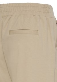 Pantalon beige avec une taille élastique et une seule poche passepoilée à l'arrière visible du côté droit.